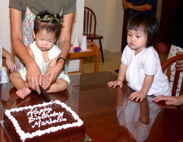 Two young children at a table with a birthday cake, one being helped by an adult to cut it